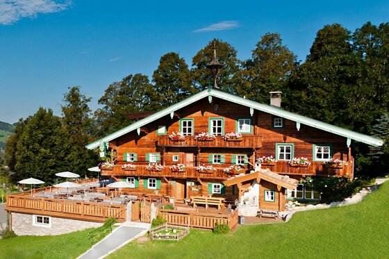 Alpiner Berggasthof aus dunklem Holz mit blumengeschmückten Balkonen und einer großen Sonnenterrasse vor Waldkulisse.