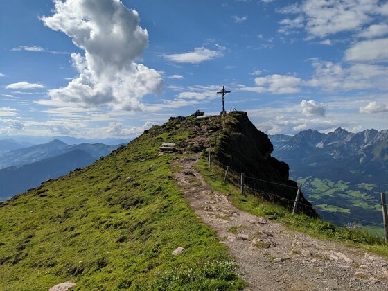 Wanderweg zum Gipfelkreuz Ein schmaler Wanderweg führt über einen grasbewachsenen Bergrücken zu einem Gipfelkreuz unter blauem Himmel.