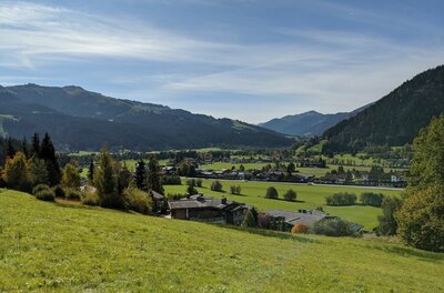 Blick von einer grünen Wiese auf ein Dorf im Tal vor bewaldeten Bergen unter blauem Himmel.