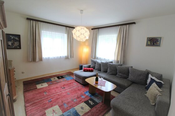 Wohnzimmer mit grauem Ecksofa, Holztisch, rotem Teppich und zwei Fenstern.