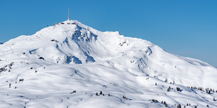 On the Kitzbüheler Horn in the Kitzbühel ski area, a snow-covered mountain landscape stretches out beneath a bright blue sky, with a lookout tower at the summit.