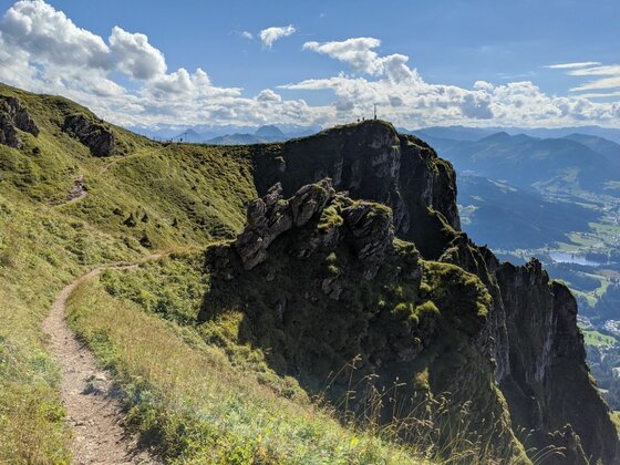 Horn Gipfel-Höhenweg Schmaler Wanderpfad an einem grasbewachsenen Berghang mit Blick auf schroffe Felsformationen und ein Gipfelkreuz.