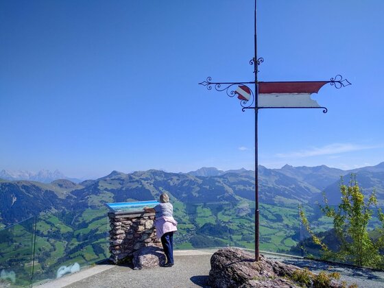 Person an einem Aussichtspunkt mit Panoramatafel und Metallkreuz vor einer Bergkulisse unter blauem Himmel.