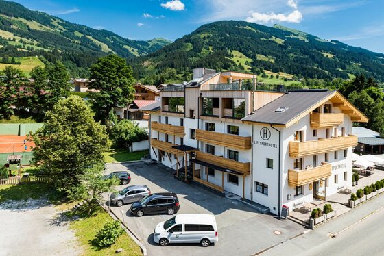 Mehrstöckiges Hotelgebäude mit Holzfronten und Parkplatz vor bewaldeter Bergkulisse.
