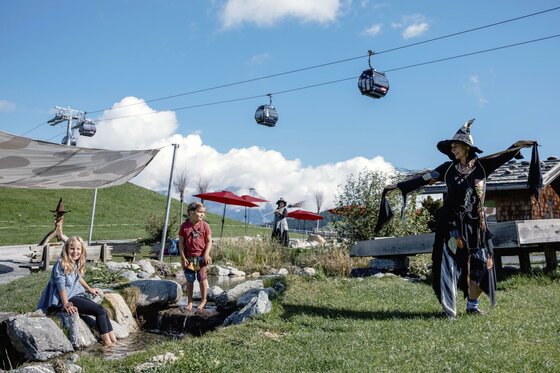 Söll_Hexenwasser_Flußlauf Kinder spielen auf einer Holzkonstruktion in einer grünen Landschaft. Eine Frau beobachtet die beiden Kinder in der natürlichen Umgebung.