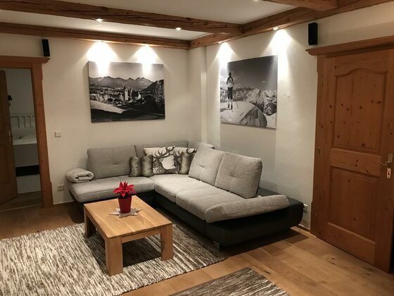 Wohnraum Apartment Hahnenkammblick Wohnzimmer mit grauem Ecksofa, Holztisch und Bergfotografien an der Wand.