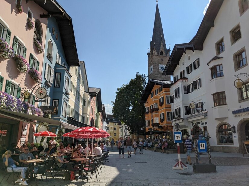 Ortszentrum von Kitzbühel Belebte Fußgängerzone mit farbenfrohen Häuserfassaden und einem spitzen Kirchturm im Hintergrund.