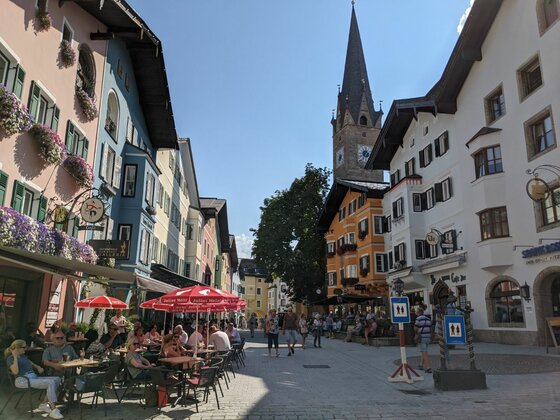 Ortszentrum von Kitzbühel Belebte Fußgängerzone mit farbenfrohen Häuserfassaden und einem spitzen Kirchturm im Hintergrund.