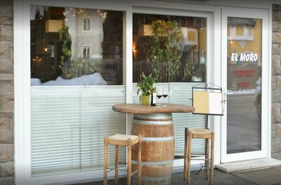 Außenbereich Pizzeria El Moro Außenansicht eines Restaurants mit einem Weinfass als Stehtisch, zwei Barhockern und Weingläsern vor einem Fenster.