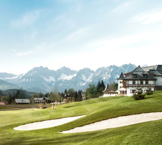 Hotelansicht mit Golfplatz Eine malerische Landschaft mit einem Golfplatz und einem eleganten Gebäude. Im Hintergrund erheben sich majestätische Berge unter einem klaren Himmel.