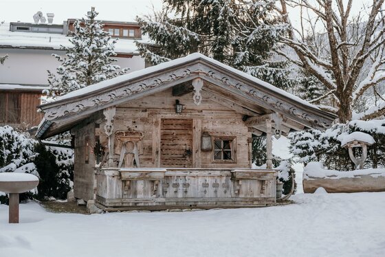 Kleine Holzhütte mit schneebedecktem Satteldach und verzierten Balken in einer Winterlandschaft.