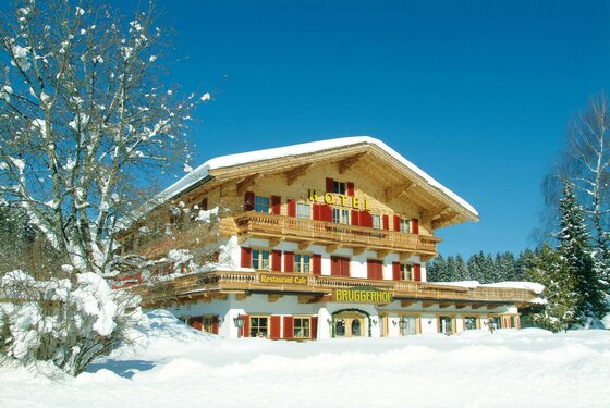 Verschneites Hotel Bruggerhof mit Holzfassade und roten Fensterläden unter blauem Himmel.