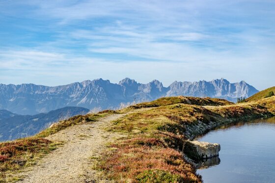 Wanderweg am Pengelstein Schotterweg auf einem Graskamm neben einem Bergsee mit Blick auf ein zerklüftetes Gebirgsmassiv unter blauem Himmel.
