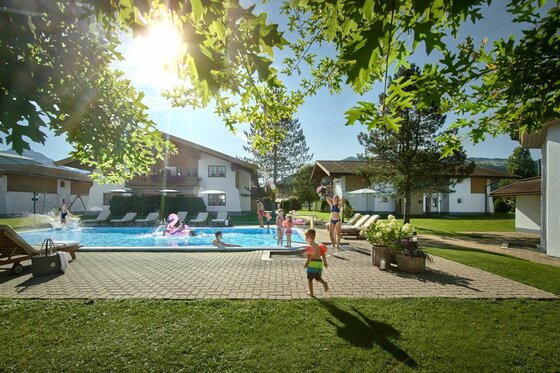 Personen in einem Außenpool vor Hotelgebäuden bei Sonnenschein.