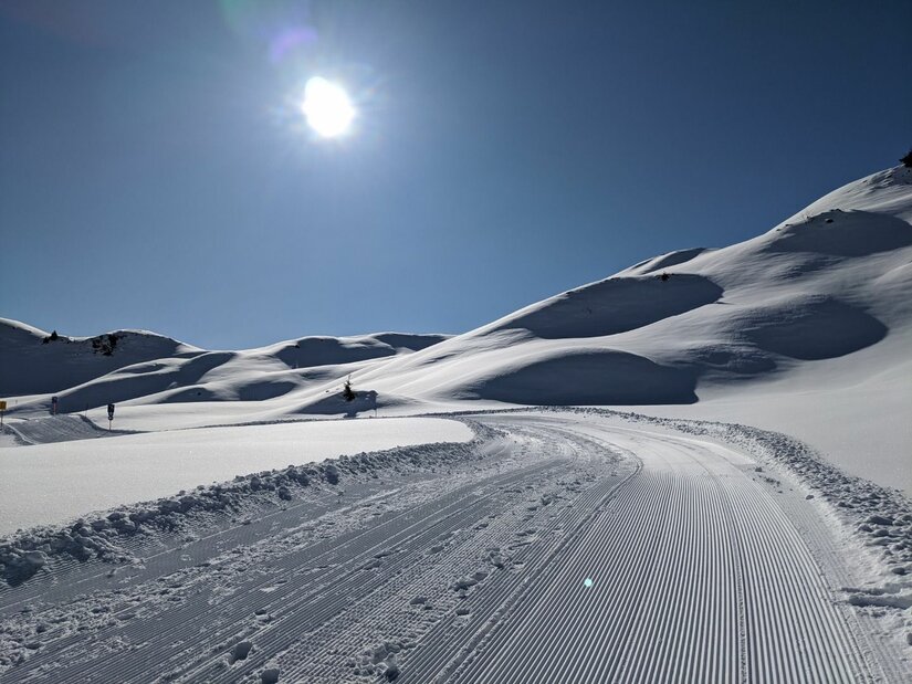 Präparierter Winterwanderweg in einer verschneiten Hügellandschaft unter blauem Himmel mit Sonnenschein.