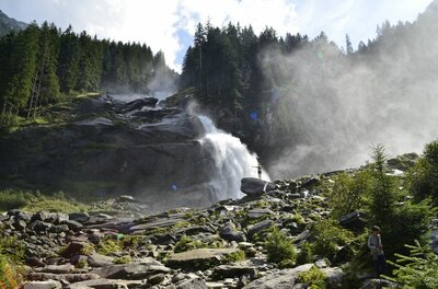 Krimmler Wasserfälle im Nationalpark Ein großer Wasserfall stürzt über dunkle Felsen in ein steiniges Becken, umgeben von Nadelwald.