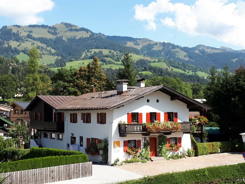Zweistöckiges weißes Alpenhaus mit roten Fensterläden und Blumenschmuck vor bewaldeten Bergen.
