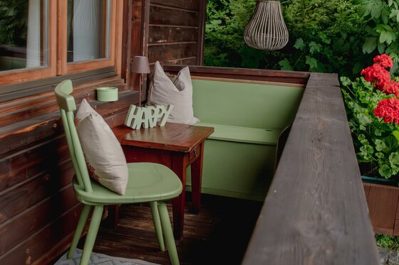 Holzbalkon mit grüner Sitzbank, einem Stuhl, kleinem Tisch und roten Blumen im Hintergrund.