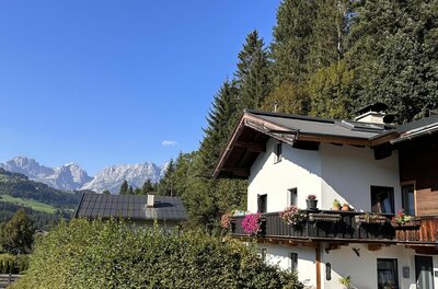 Alpines Wohnhaus mit Blumenbalkon vor bewaldetem Hang und Gebirgskette unter blauem Himmel.