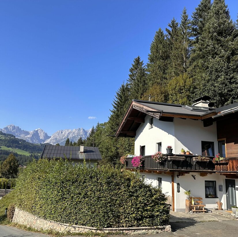 Alpines Wohnhaus mit Blumenbalkon vor bewaldetem Hang und Gebirgskette unter blauem Himmel.