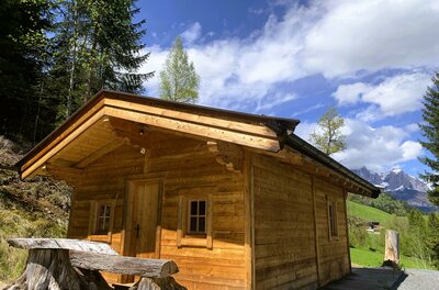image00001 Eine gemütliche Holzhütte umgeben von Bäumen und einer malerischen Landschaft. Der Himmel ist blau mit einigen Wolken.