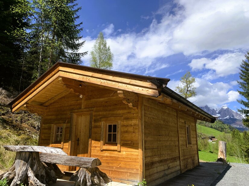 Eine gemütliche Holzhütte umgeben von Bäumen und einer malerischen Landschaft. Der Himmel ist blau mit einigen Wolken.