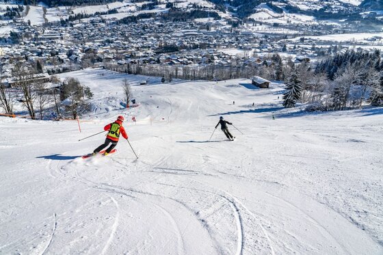 Skifahren am Hahnenkamm Zwei Skifahrer auf einer präparierten Piste oberhalb eines verschneiten Ortes im Tal.