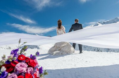 Ein Brautpaar steht auf einem schneebedeckten Berg mit atemberaubender Aussicht. Im Vordergrund sind bunte Blumen zu sehen.