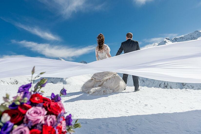 Ein Brautpaar steht auf einem schneebedeckten Berg mit atemberaubender Aussicht. Im Vordergrund sind bunte Blumen zu sehen.