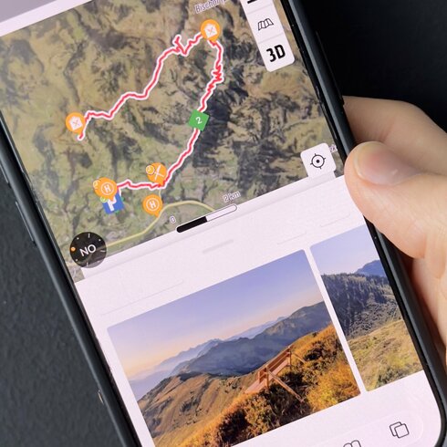 A hand is holding a smartphone displaying a detailed map of hiking trails in the mountains, surrounded by nature.