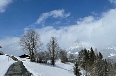 Geräumter Asphaltweg in einer verschneiten Alpenlandschaft mit Blick auf einen wolkenverhangenen Berggipfel.