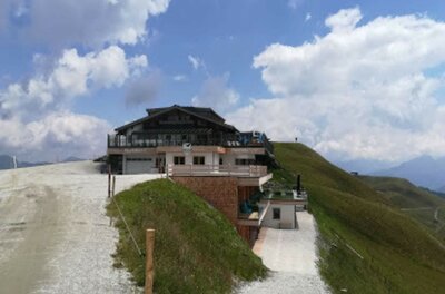 Ehrenbachgraben im Sommer Berghütte mit Holzterrasse an einem Schotterweg vor grasbewachsenen Hängen unter bewölktem Himmel.
