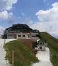 Berghütte mit Holzterrasse an einem Schotterweg vor grasbewachsenen Hängen unter bewölktem Himmel.