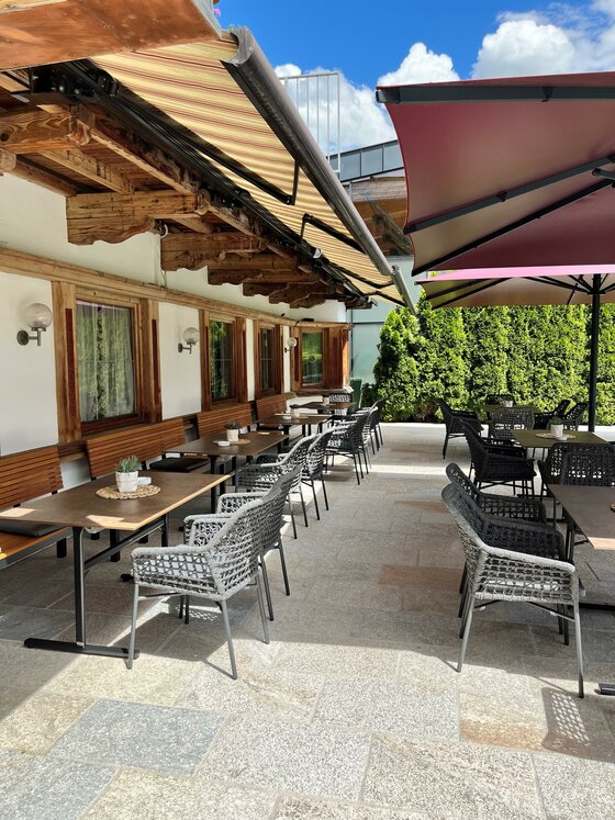 Terrasse des Bruggerhofs Außenterrasse mit Holztischen, grauen Korbstühlen und Sonnenschirmen vor einer weißen Hauswand.