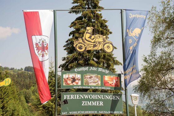 Gasthof Alte Wacht Jochberg Schild des Gasthofs Alte Wacht mit Motorrad-Silhouette, Tiroler Flagge und blauer Fahne vor Bäumen.