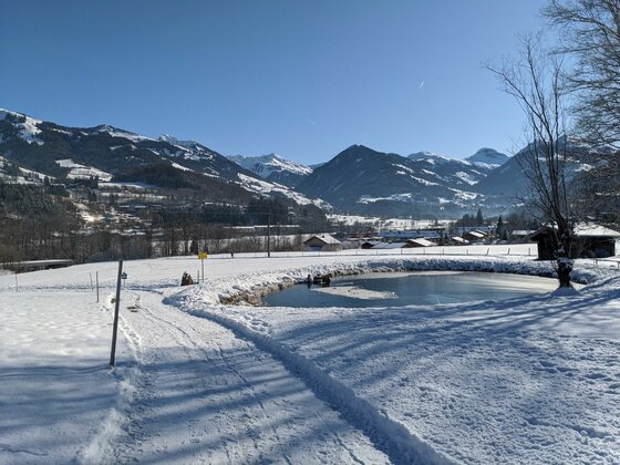 Verschneite Landschaft mit einem kleinen Teich, einem Winterwanderweg und schneebedeckten Bergen im Hintergrund.