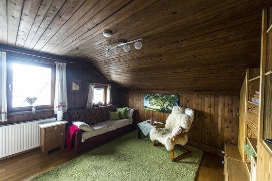 App Wilder Kaiser Wohnzimmer Wohnbereich mit Holzverkleidung, Sofa, Sessel mit Fellauflage und grünem Teppich unter einer Dachschräge.