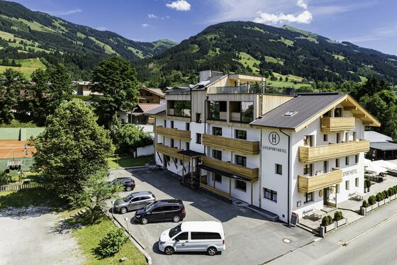 Mehrstöckiges Hotelgebäude mit Holzfassadenelementen, Parkplatz und bewaldeten Bergen im Hintergrund.