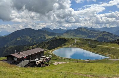 Berghütte neben einem runden Speichersee in einer alpinen Graslandschaft mit Bergpanorama im Hintergrund.