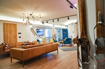 Lounge im Safestay Kitzbühel Moderner Loungebereich mit braunem Ledersofa, bunten Sesseln, Holzboden und dekorativen Skiern an der Wand.