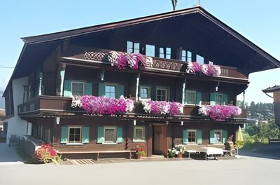 Ein traditionelles altes Haus mit mehreren Stockwerken und vielen bunten Blumen auf den Balkonen. Der Himmel ist klar und die Umgebung ist grün.