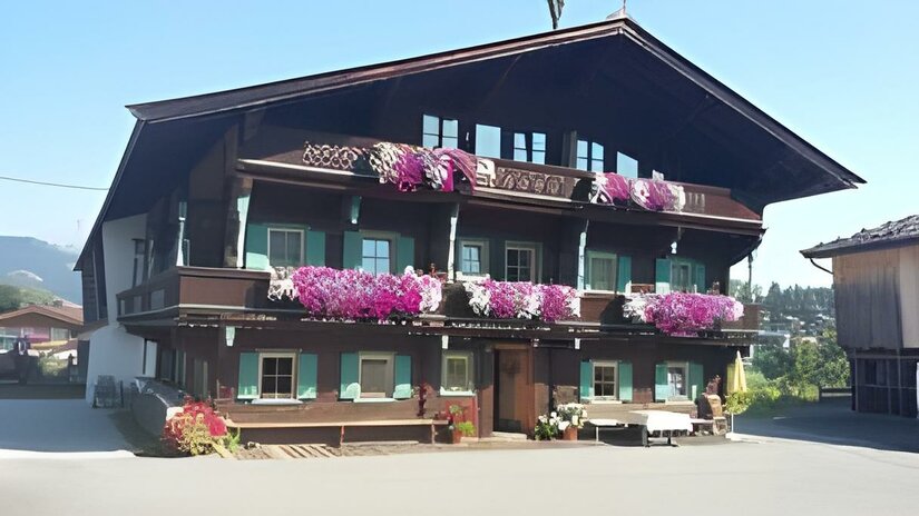 Hölzlhof-Vermietung-Bauernhof-Kitzbühel-Haus Ein traditionelles altes Haus mit mehreren Stockwerken und vielen bunten Blumen auf den Balkonen. Der Himmel ist klar und die Umgebung ist grün.
