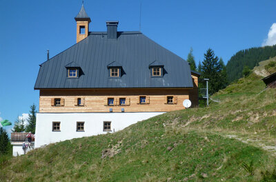 Dreistöckiges alpines Gebäude mit Holzfassade, weißem Erdgeschoss und dunklem Satteldach vor blauem Himmel.