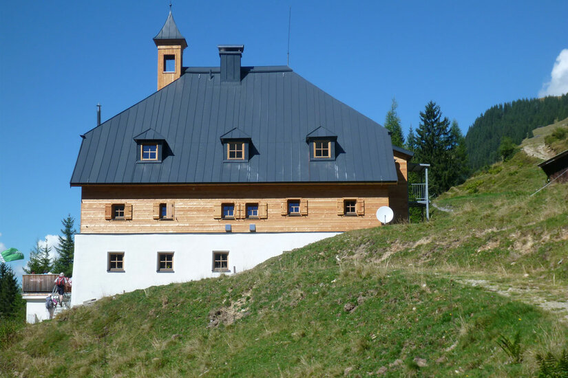 Kelchalm - Bochumer Hütte Dreistöckiges alpines Gebäude mit Holzfassade, weißem Erdgeschoss und dunklem Satteldach vor blauem Himmel.