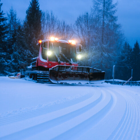 In Kitzbühel, a red snow groomer is preparing the cross-country ski trail in a snow-covered landscape dotted with fir trees in the twilight.