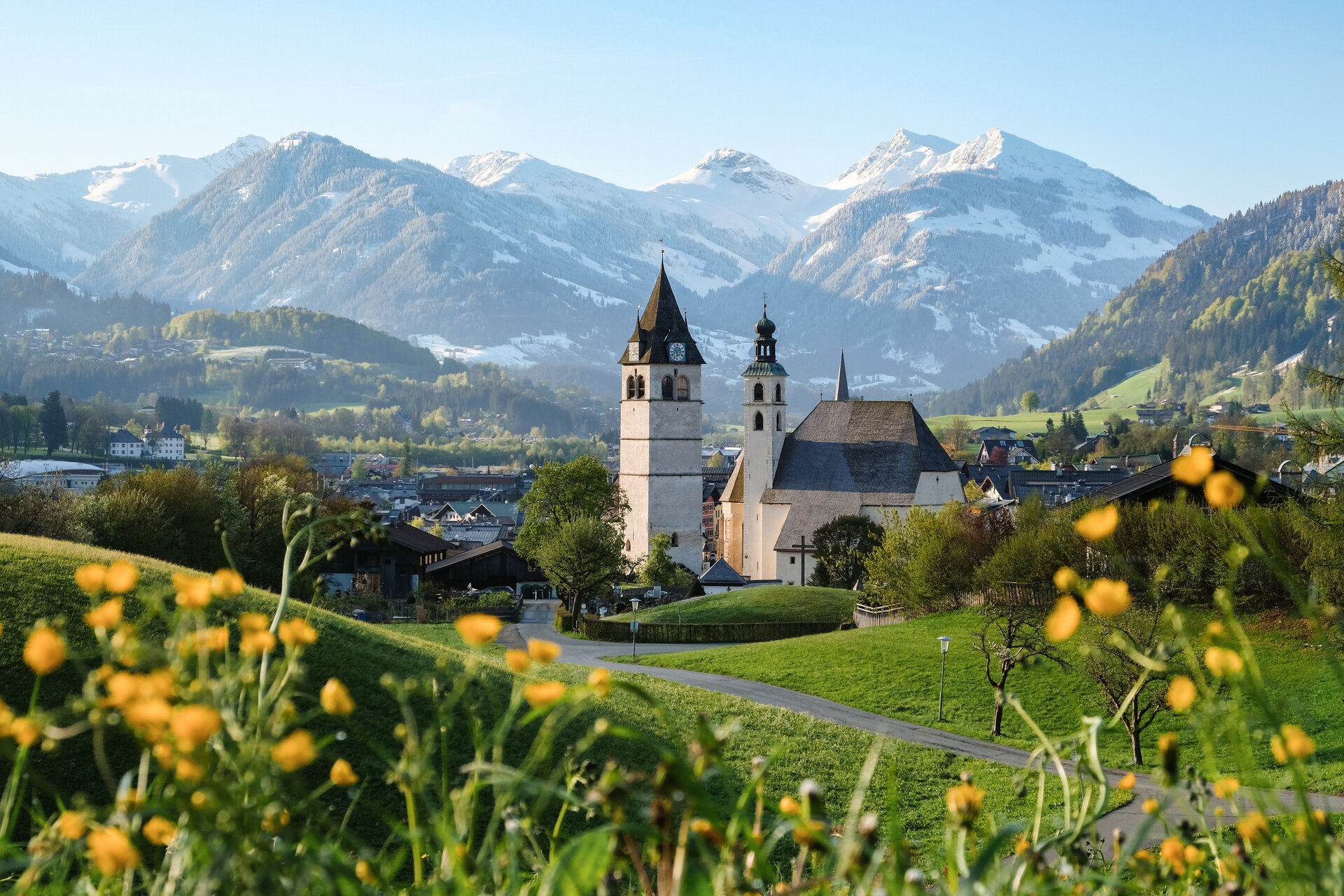 Eine malerische Landschaft mit einer Kirche im Vordergrund, umgeben von blühenden Wiesen und majestätischen Bergen im Hintergrund.