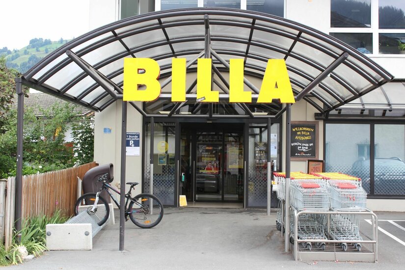 Eingangsbereich eines Billa Supermarktes mit gelbem Schriftzug, Glasvordach, Einkaufswagen und einem Fahrradständer.