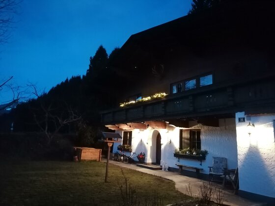 Ansicht seitlich mit Beleuchtung Beleuchtetes alpines Wohnhaus in der Dämmerung mit Balkon und Gartenbereich.