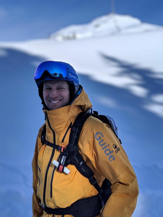 Bergführer Powderflo-Florian Ein Bergführer in gelber Jacke mit Helm und Lawinenrucksack vor einer verschneiten Bergkulisse.