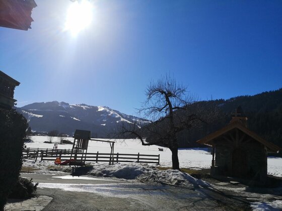 Winterlandschaft am Griesbach Verschneite Berglandschaft mit kleiner Kapelle, Baum und Spielplatz unter blauem Himmel bei Sonnenschein.